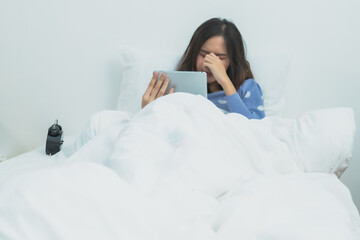 Tired young Asian woman feeling eye strain or headache while using digital tablet in bed, stressed girl suffering from digital fatigue and exhaustion in cozy bright bedroom