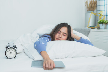Exhausted young Asian woman sleeping on white bed while holding a digital tablet, tired girl falling asleep in cozy bedroom with alarm clock, sleep deprivation and technology addiction concept.