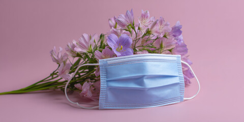 A blue surgical mask placed in front of a bouquet of delicate purple flowers, showcasing a blend of health and beauty on a pink background.