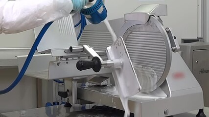 In a professional kitchen, a worker wearing protective clothing meticulously cleans a slicer machine. The process involves spraying the machine with a cleaning solution and scrubbing it thoroughly.