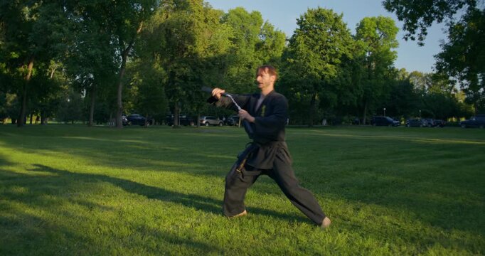 A male karateka with nunchaku trains outdoors.. A male karateka trains on the grass with nunchaku and tonfa.