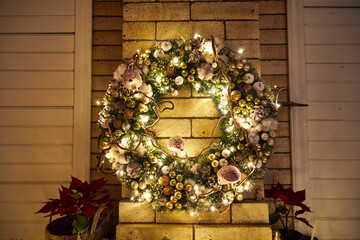 Beautiful Christmas wreath decorated with lights, ornaments and winter greenery hanging on a brick wall. Warm festive holiday atmosphere, cozy seasonal home decor and magical Christmas mood
