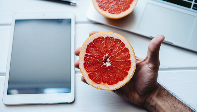 Hand holding a vibrant grapefruit half over a white desk with a tablet and laptop.