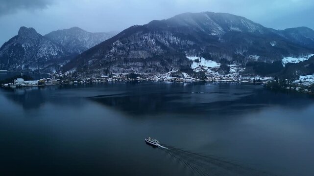 Aerial drone flying sideways behind boat on Traunsee lake with winter alpine landscape, Austria