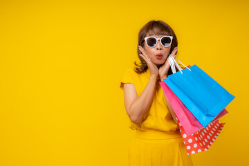 Cheerful young Asian woman in vibrant yellow jumpsuit and straw hat holding two colorful ice cream cones, smiling at camera on isolated yellow background, summer vacation and refreshment concept.
