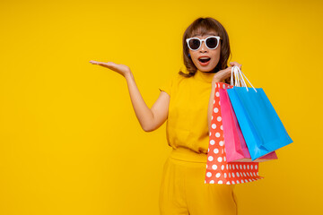 Cheerful young Asian woman in vibrant yellow jumpsuit and straw hat holding two colorful ice cream cones, smiling at camera on isolated yellow background, summer vacation and refreshment concept.