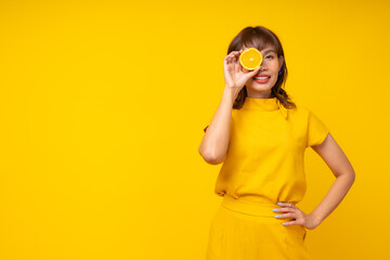 Playful Happy Asian Woman Holding Fresh Orange Slice Over Eye on Vibrant Yellow Background, Summer Health and Vitamin C Beauty Skincare Concept Studio Portrait.