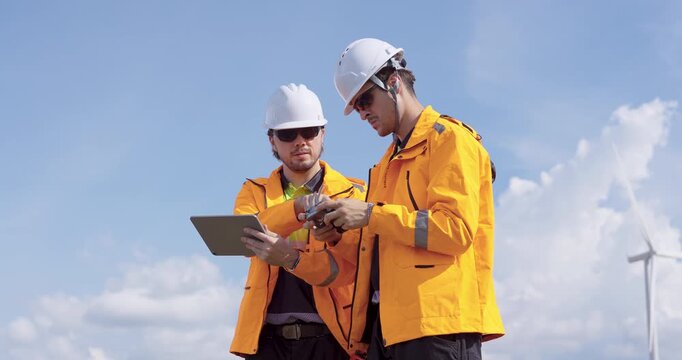 Two Caucasian engineers wearing orange safety jacket operate drone to inspect wind turbine performance using tablet for data recording maintaining renewable energy system with teamwork and precision