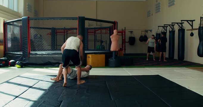 Two mixed martial artists practice takedowns on sports mats in a training hall.