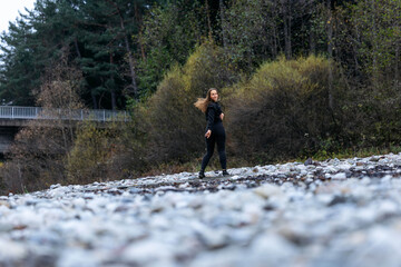 Obraz premium Young woman with long hair wearing black outfit is walking along rocky riverbank, surrounded by trees and autumn foliage, capturing a moment of tranquility in nature