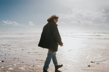 A woman is walking on the beach wearing a black coat. The sky is cloudy and the sun is shining