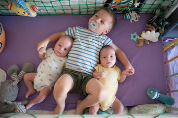 Three young children are in a crib. An older brother with a pacifier holds hands with his two infant twin siblings on a purple mattress surrounded by toys