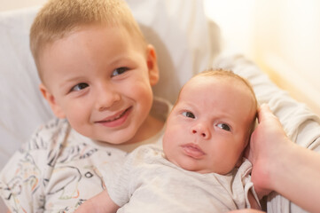 A young boy with blond hair smiles as he holds his newborn brother who looks curiously at the camera. They are both in light clothing and seem comfortable and loved