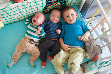 Three brothers lie next to each other in their crib on a light blue sheet. One brother is crying, while the other two are looking content and smiling at the camera