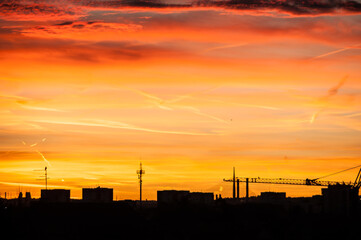 A vibrant sunset fills the sky with orange and red colors. Below, a silhouetted cityscape including buildings, antennas, and construction cranes stands against the colorful twilight