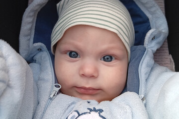 A baby with blue eyes looks directly into the camera while sitting in a stroller. The baby wears a light blue outfit and a striped hat