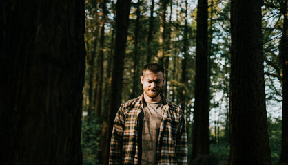 A man is standing in a forest, looking at the camera. He is wearing a plaid shirt and he is in a contemplative mood