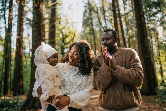 A family of three, a man and two children, are walking through a forest. The man is holding a baby and the other child is wearing a white jacket. The family is smiling and enjoying their time together