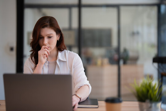 Young professional businesswoman concentrating and analyzing data on her laptop, engaging in deep thought while working remotely from a contemporary office space