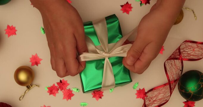 Overhead view of woman tying white silk ribbon on green gift box. Top down shot female packing present at white background. Overhead flat lay of Christmas art and craft equipment surrounding 