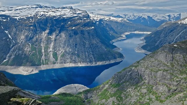 A tourist in a bright yellow jacket stands with arms outstretched on the narrow rock ledge of Trolltunga. Steep cliffs, snow-patched mountains, and dramatic alpine terrain.