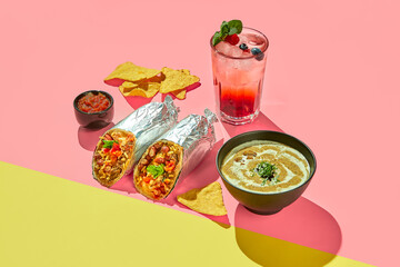 Mexican cuisine dishes, set meal of burrito, mushroom soup, corn chips nachos and berry lemonade on colored background