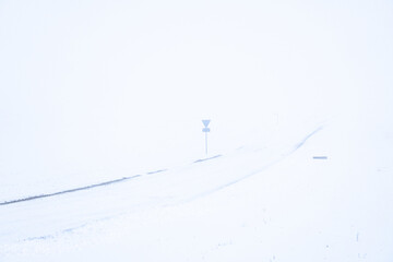 Minimalist winter road disappearing into dense fog and snowfall, empty rural landscape with road sign barely visible, expressing isolation, uncertainty and silent Nordic seasonal mood