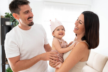 Fototapeta premium Happy family couple celebrating baby's first birthday at home, parents holding and laughing with their adorable one year old daughter wearing a crown, enjoying togetherness and love