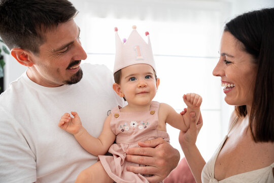 Loving parents smiling and holding their cute baby girl, celebrating her first year of life, with the child wearing a sweet party crown marking her one-year milestone