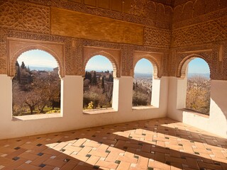 alhambra - Sunlight floods through Moorish arched windows into an ornate hall, casting long shadows on the tiled floor. The view reveals a sunny hilly landscape.