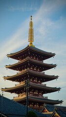 A majestic five-story red pagoda with a golden spire towers into the blue sky, surrounded by traditional Japanese temple architecture.