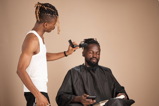 A barber gently works on a seated Black man&rsquo;s hair using a comb and brush while the client relaxes with eyes closed, holding a smartphone and an open book. The studio scene reflects a calm, modern