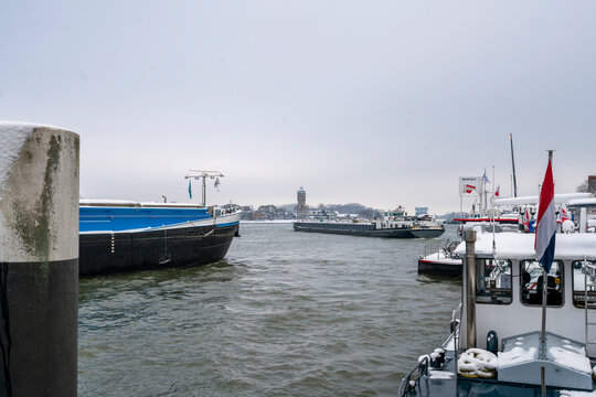 Dordrecht Netherlands - 5 January 2026: Busy winter harbour with cargo barge passing Dordrecht on a calm, snowy urban industrial waterfront maritime transport scene on thMaas River in the Netherlands.