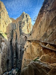 Caminito del Rey A narrow wooden walkway clings to the steep rock walls of a deep gorge. The spectacular hiking trail leads through a breathtaking rocky landscape under a blue sky.