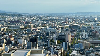 A wide panoramic view over over Kyoto a dense urban cityscape in Japan with countless buildings, a long train line, and construction cranes in daylight.