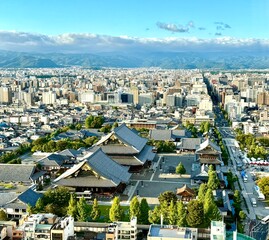 A breathtaking view over the city of Kyoto shows the massive grey roofs of a traditional temple complex amidst modern buildings against a distant mountain range.