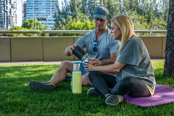 Middle aged couple sharing traditional mate infusion while resting on the grass after a workout