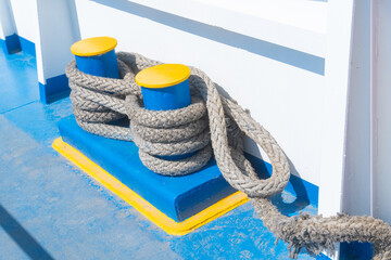 A detailed view of a thick rope tied around a blue bollard, located on a boat near a calm water body. Represents maritime activities, boating and nautical themes © Elena