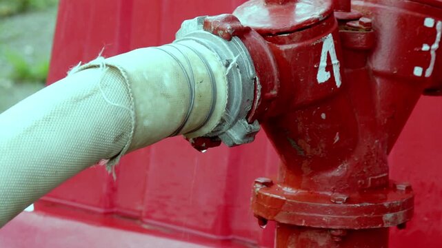 Fire hydrant is connected to fabric hose with metal coupling leaking water. Red hydrant body with painted marking visible.