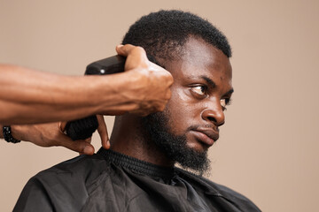 Close-up of a professional barber using electric clippers to trim the side of an adult Black man’s hair during a grooming session. The client wears a black barber cape, showing focused detail 