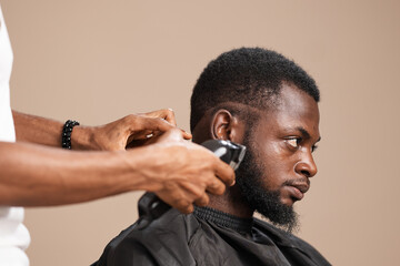 Side profile of an adult Black man wearing a barber cape while a professional barber trims the back and sides of his hair with electric clippers during a grooming session in a studio with a neutral 