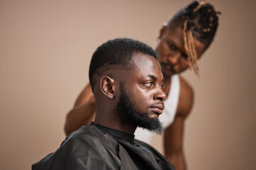 Side profile portrait of an adult Black man with a fresh low fade haircut and full beard wearing a barber cape, while a barber stands behind him during a professional grooming session in a studio with © The Yudel Media