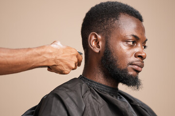 Close-up side profile of an adult Black man with short natural hair and a shaped beard wearing a barber cape while a barber trims the back of his head during a professional grooming session 