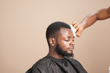 Side profile of an adult Black man with short natural hair and a groomed beard wearing a barber cape while a barber gently wipes his forehead during a professional grooming session in a studio setting