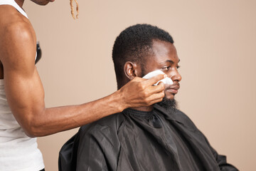 Close-up side profile of an adult Black man with short natural hair and a shaped beard wearing a barber cape, seated during a professional grooming session against a neutral studio background.
