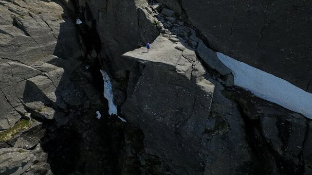 Tourist standing on a narrow rock crevice at Trolltunga in Norway. Close aerial view highlighting dramatic geology and raw mountain landscape in bright daylight.