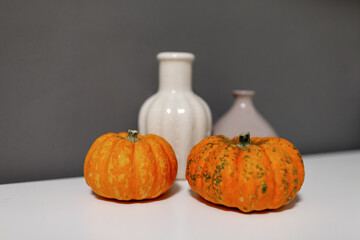 Two vibrant orange pumpkins placed on a white surface, accompanied by elegant ceramic vases, creating a cozy autumn atmosphere with rich textures and warm colors
