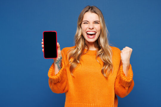 Young happy excited woman wear yellow sweater casual clothes hold in hand use mobile cell phone with blank screen workspace area isolated on plain blue background studio portrait. Lifestyle concept.