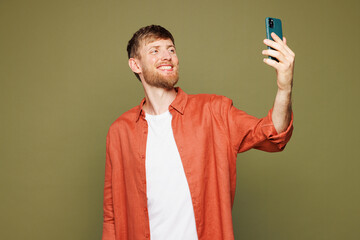 Young smiling happy man wear basic white t-shirt orange shirt hold in hand use doing selfie shot on mobile cell phone post photo on social network isolated on plain green background studio portrait