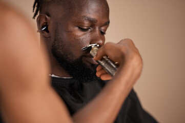 A close-up of an adult African man with a beard wearing a haircut cape and wireless earbuds as a barber trims his moustache area with a detail trimmer in a studio.